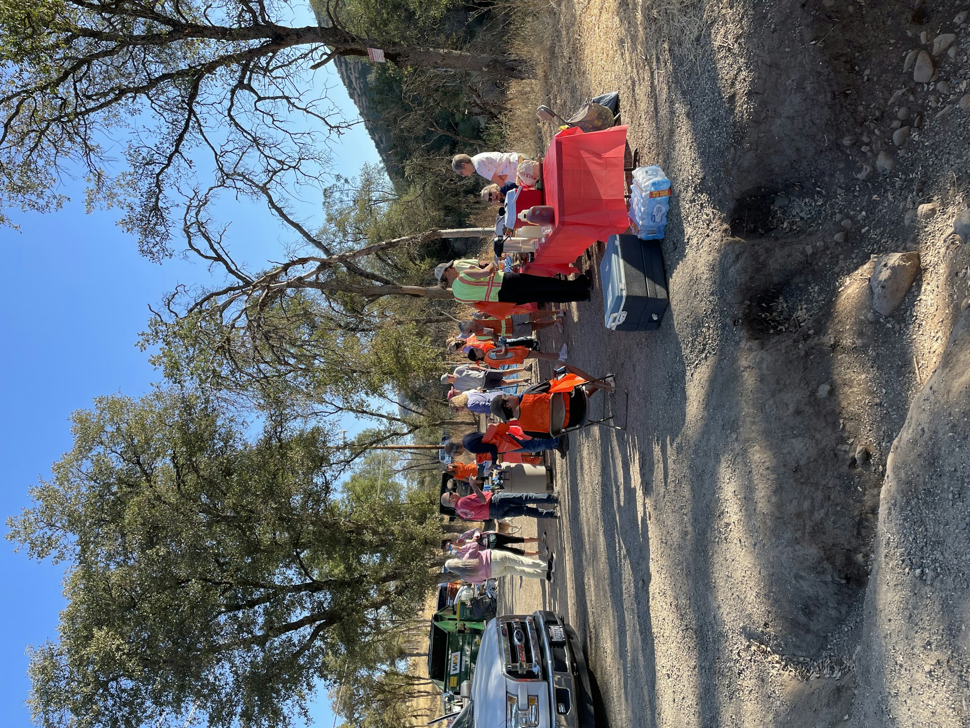 A group of people in orange vests gathered around tables outdoors, with trees and a clear sky in the background.