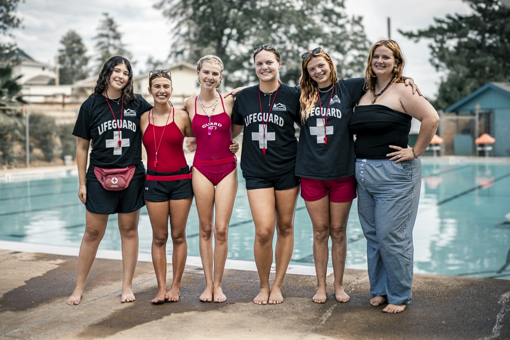 Six people, some in lifeguard attire, standing together by a pool, smiling.