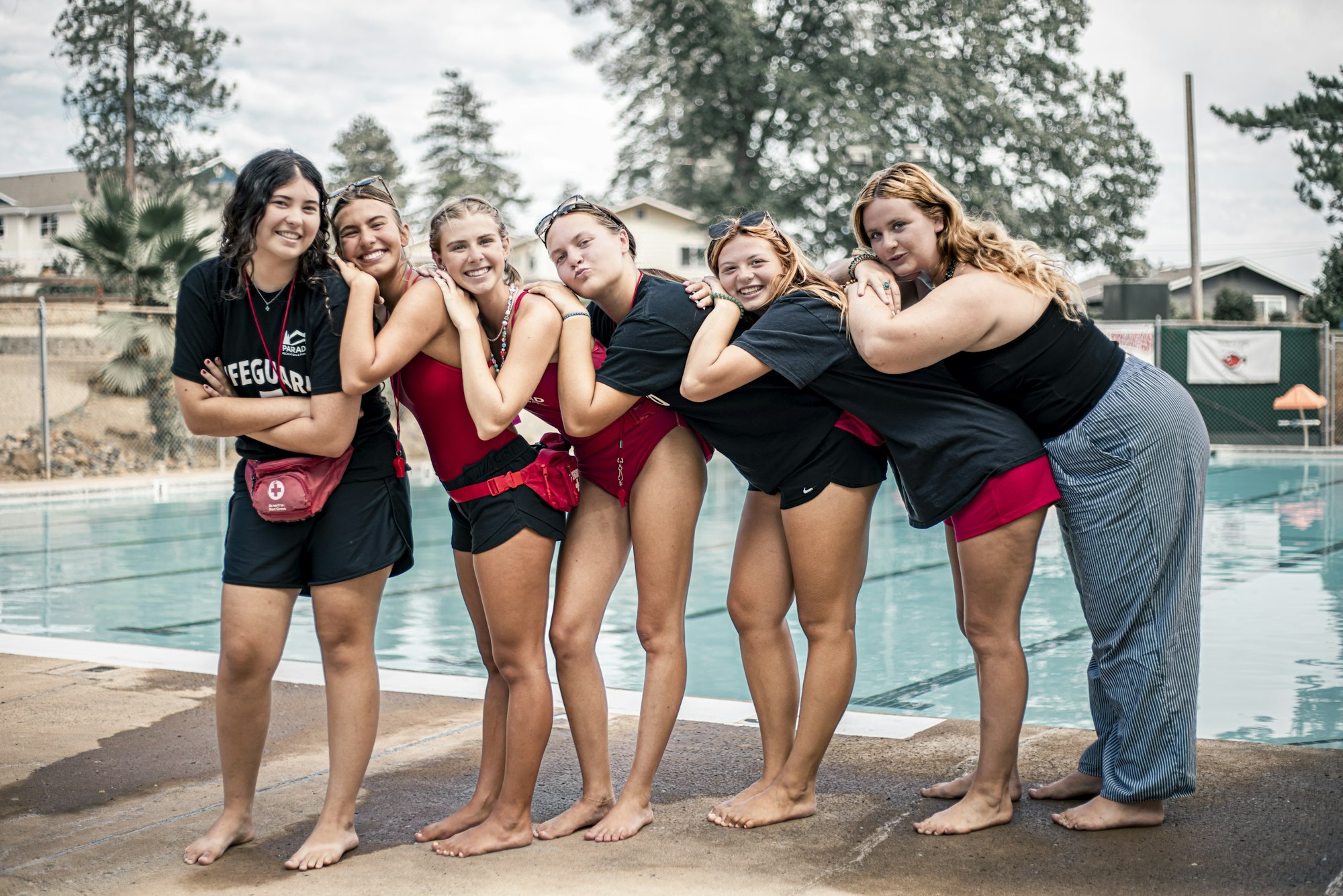 Six smiling people in lifeguard attire pose playfully by a pool.