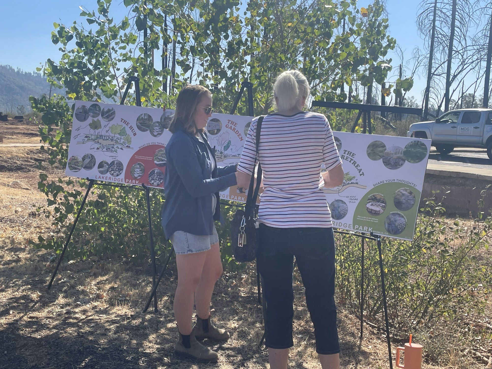Two people stand outdoors in front of an informational display board featuring diagrams and images about vegetation or land restoration. One is pointing at the board while the other listens. The scene takes place on a sunny day, with trees and a road in the background.