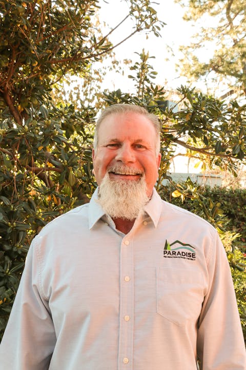 A man with a white beard smiling, wearing a shirt with "Paradise Recreation & Park District" logo, standing before greenery.