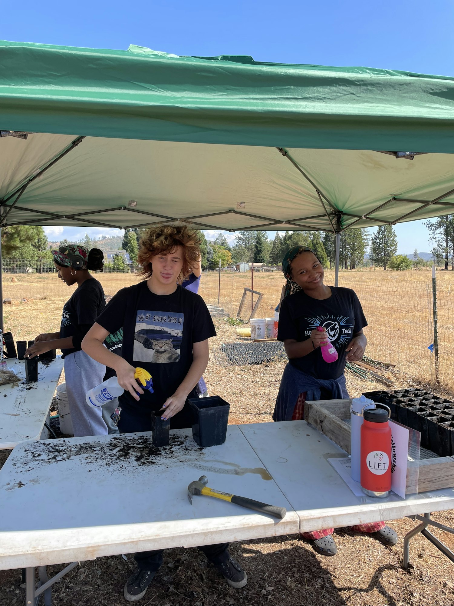 Two young individuals are working under a green canopy, engaged in planting activities with tools and supplies on a table.