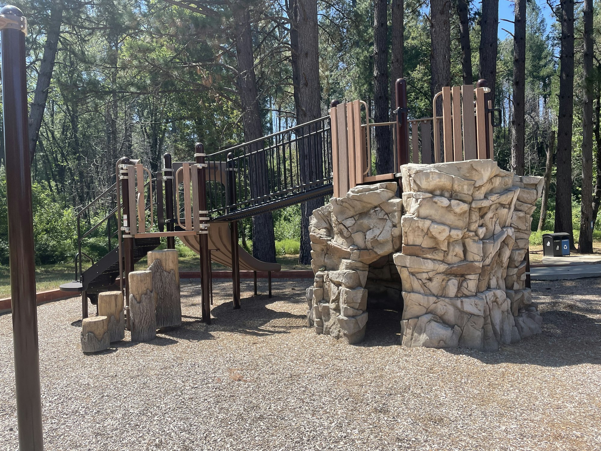 A playground with a climbing structure, slide, and artificial rock in a wooded area.