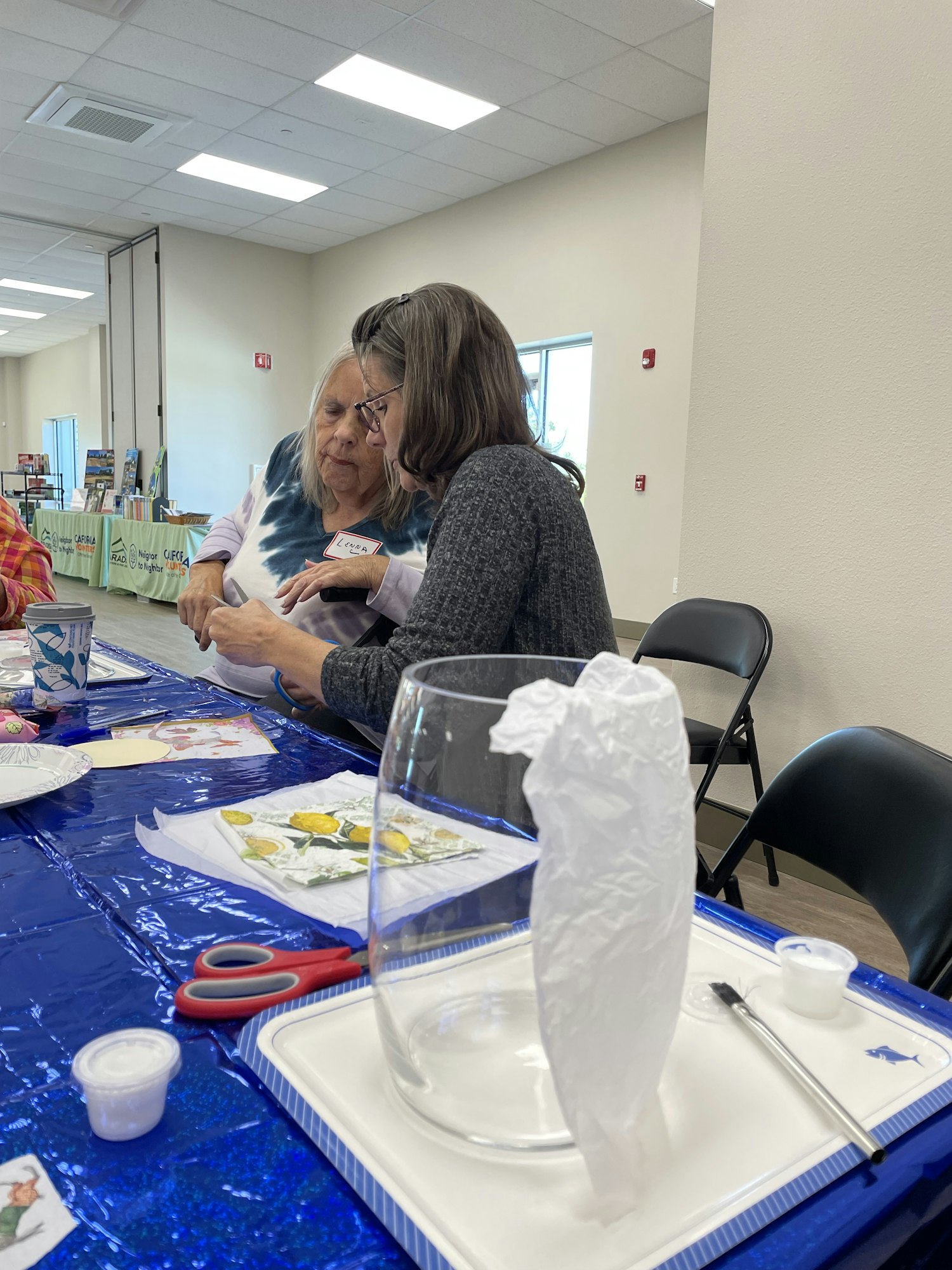 Two women are engaged in an art or crafting activity at a table, with various supplies and a colorful table covering.