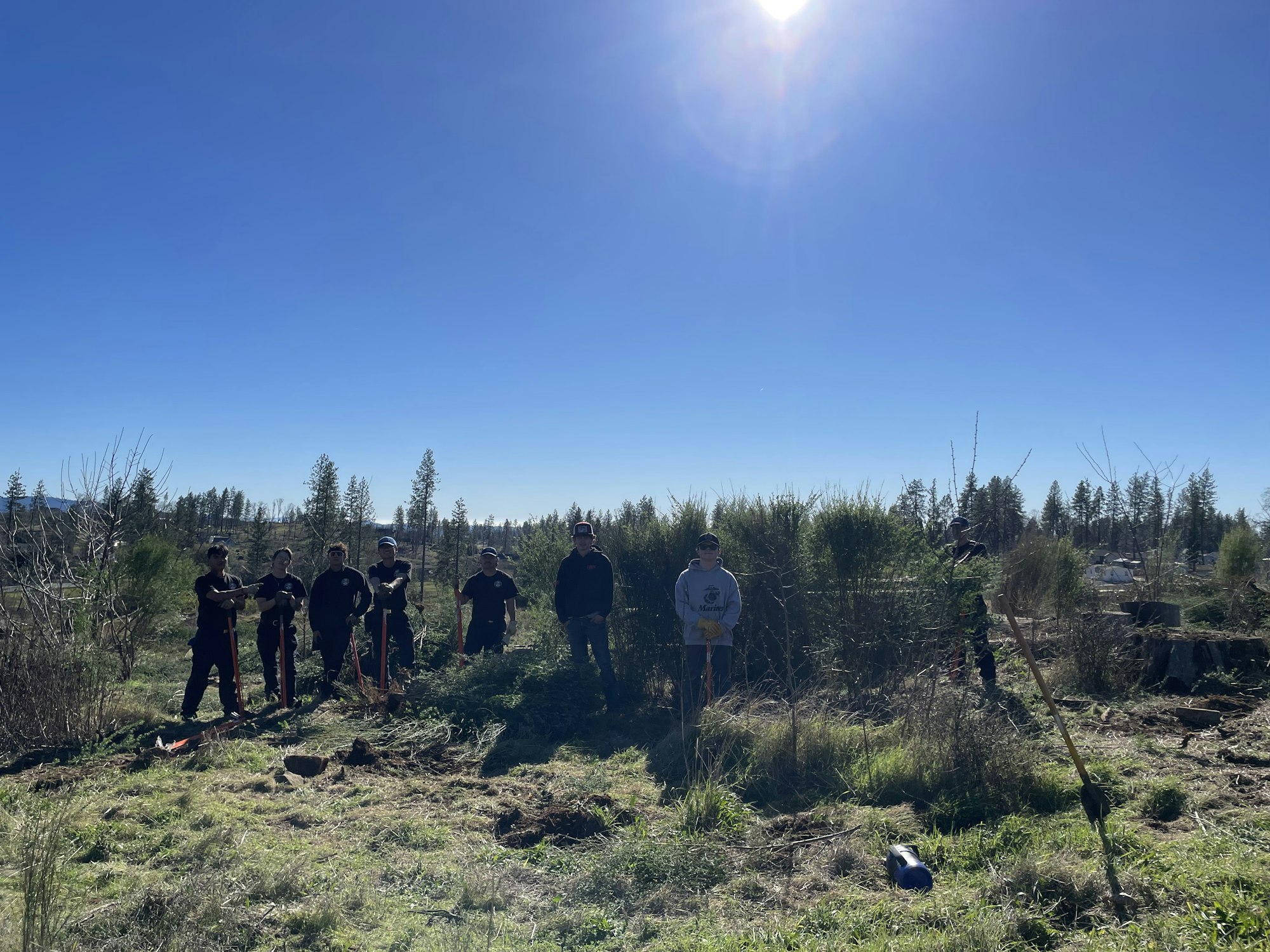 A group of people stands outdoors, equipped with tools, in a clearing with trees and blue skies in the background.