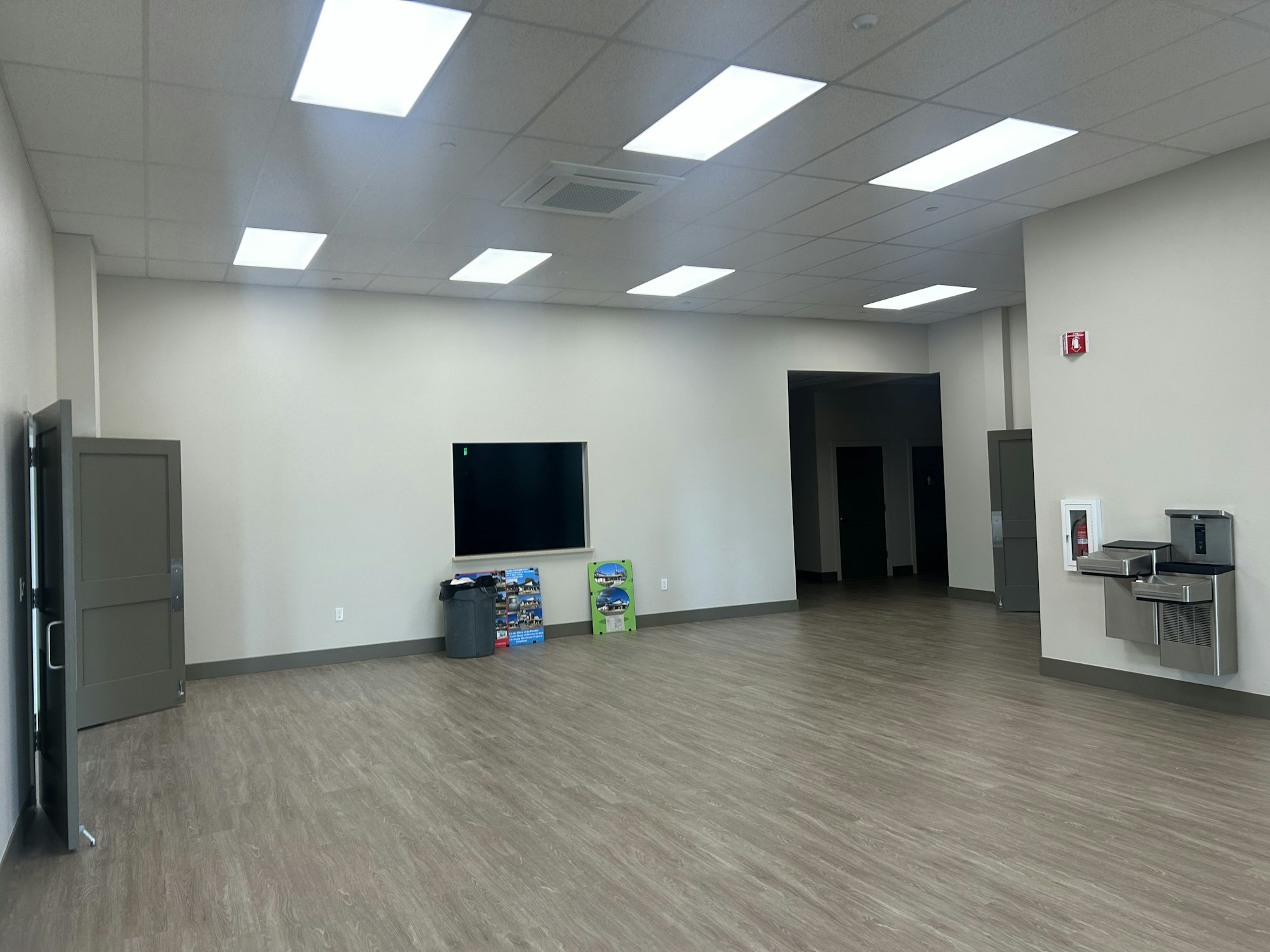 A large room with wooden flooring, a TV, some boxes, a water fountain, and ceiling lights.