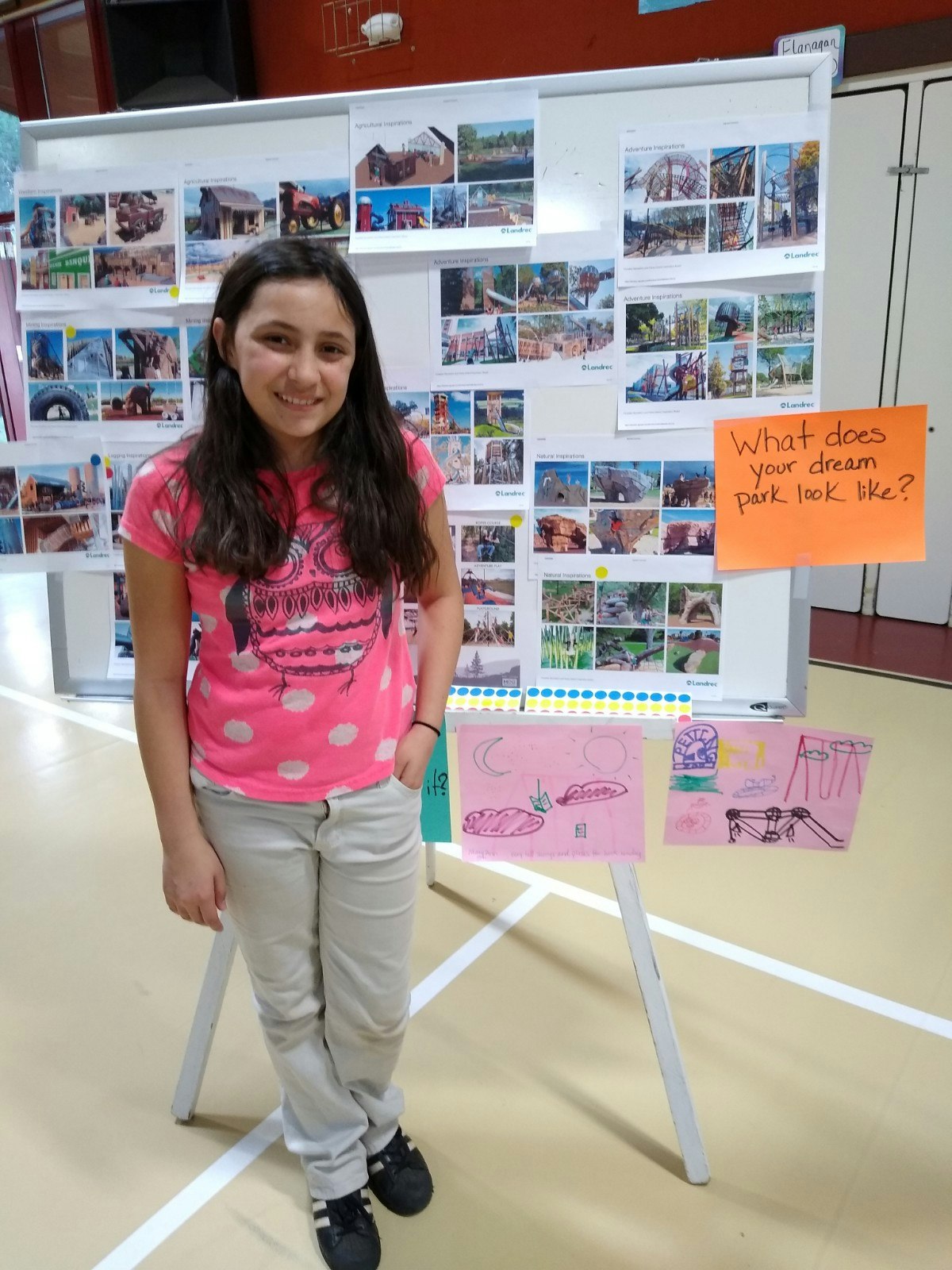 A young girl stands beside her project about dream parks, with images pinned behind her and her drawings on display.