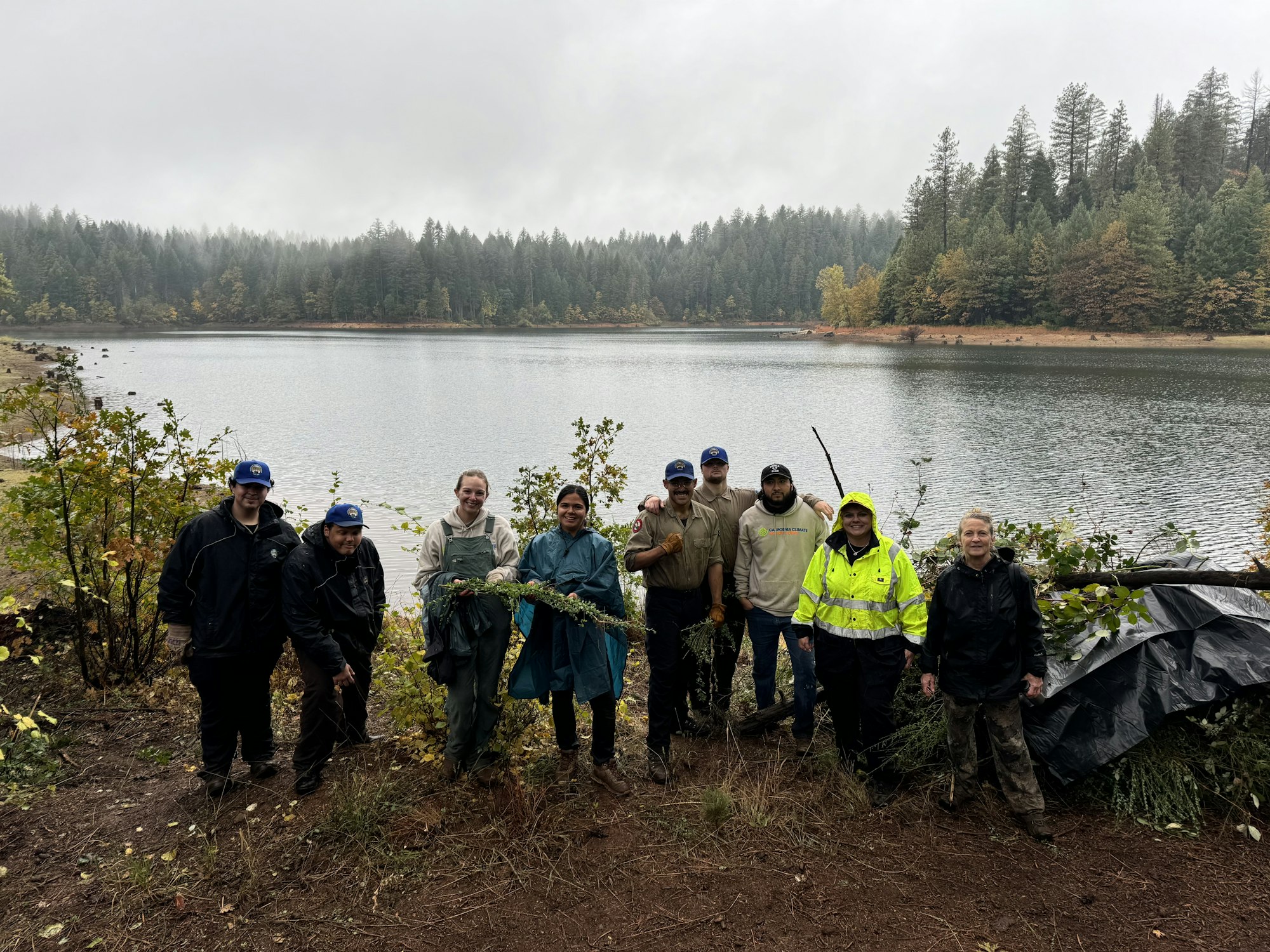A group of people stands by a lake in misty weather, surrounded by trees, possibly involved in a conservation or cleanup effort.