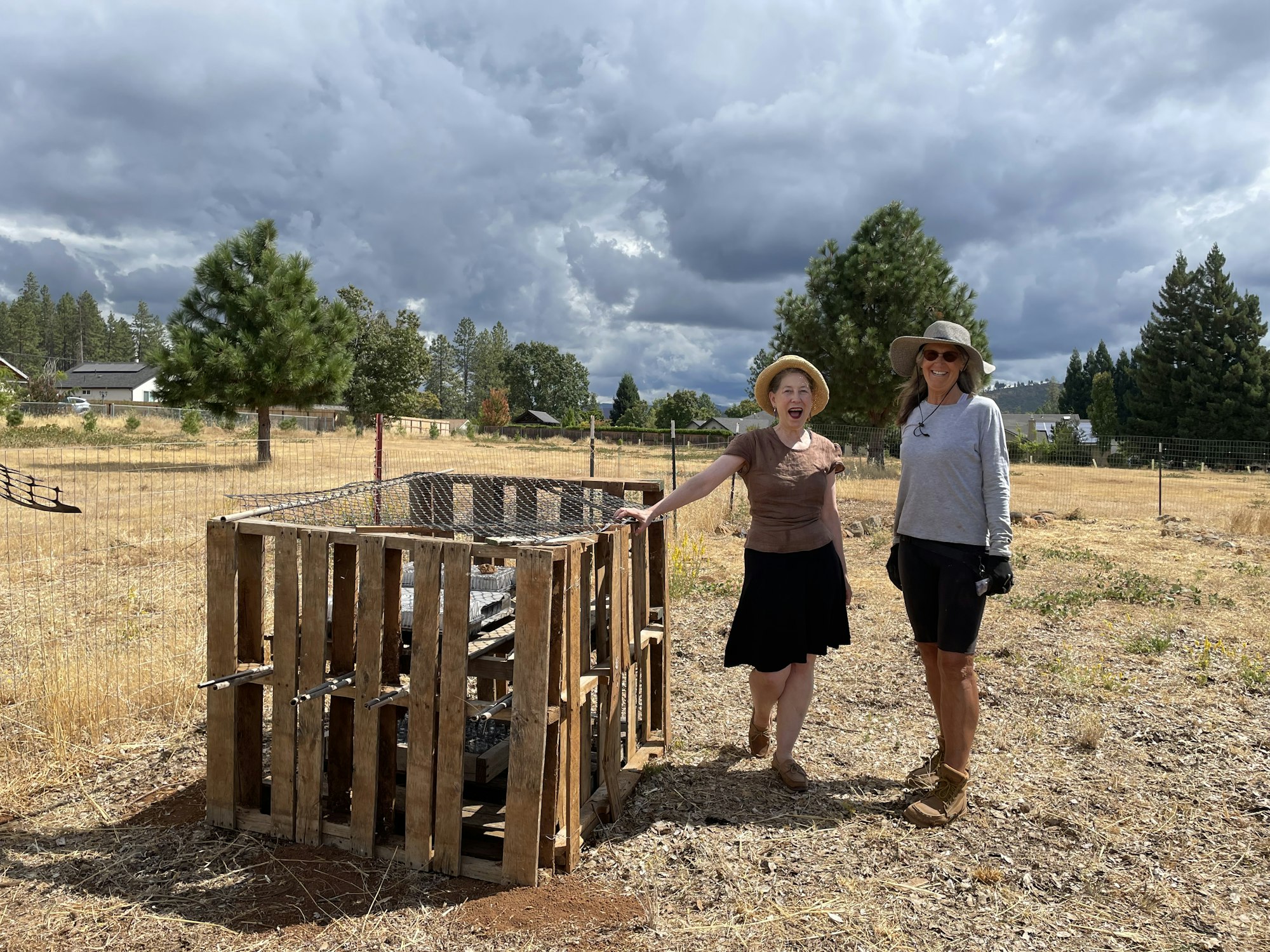 Two women stand beside a wooden compost bin in an open, grassy area under a cloudy sky, smiling and enjoying their time outdoors.