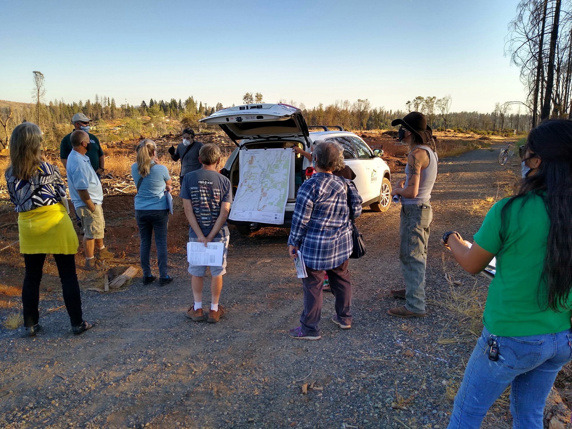A group of people gathers outdoors, discussing a map near a car in a deforested area, likely related to land management or recovery efforts.