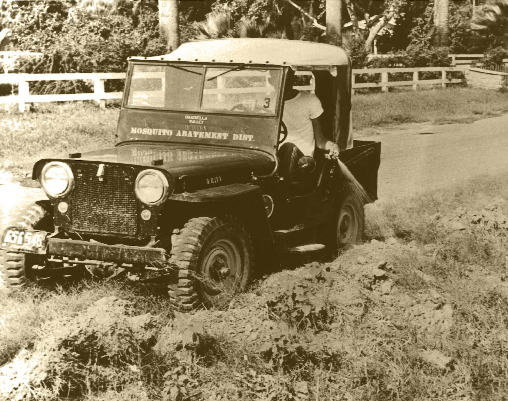 A vintage jeep labeled "Mosquito Abatement Dist." sprays pesticide while driving on a rural road.