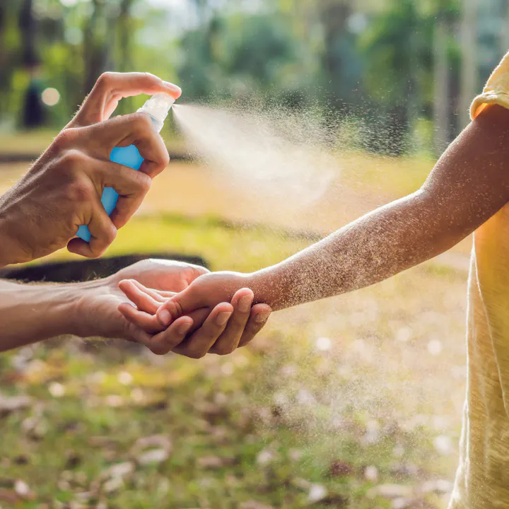 A person is spraying a liquid from a bottle onto another person's arm in a sunny outdoor setting.