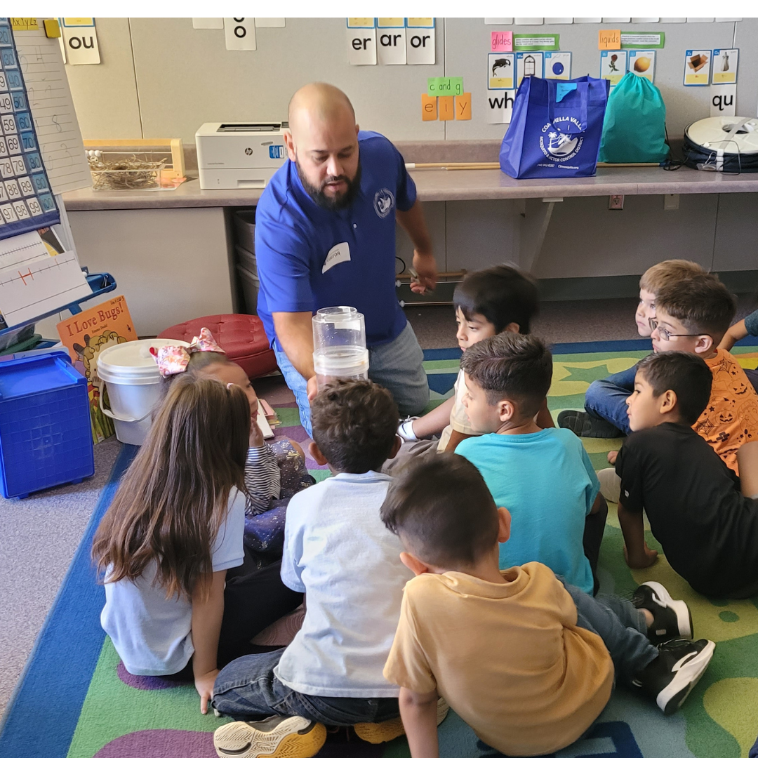 A teacher is demonstrating something to a group of attentive children in a classroom setting.
