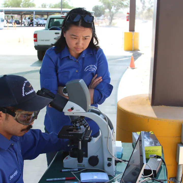 A man uses a microscope while a woman observes intently, both in blue shirts, outdoors with a vehicle in the background.