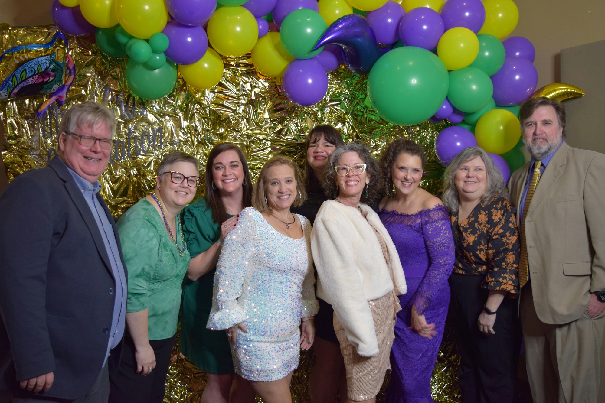 A festive group photo featuring nine people smiling, dressed elegantly, with colorful balloons in the background.