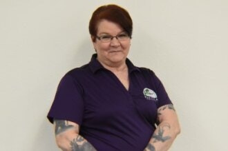 A person with tattoos wearing a purple shirt, smiling, arms crossed, standing against a white wall.