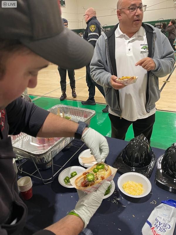 A person prepares a hot dog with toppings, while another man holds a plate, in a gym setting with others nearby.
