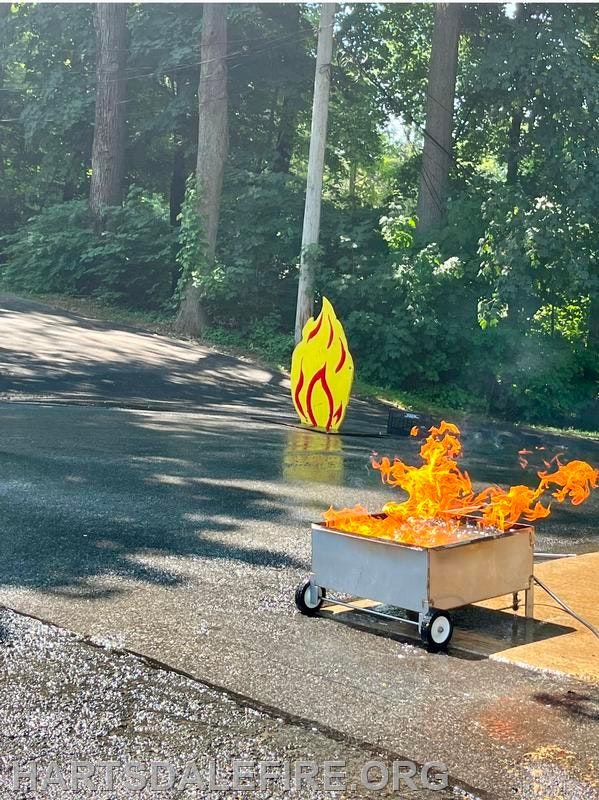 A fire training setup with a controlled flame in a metal container and a fire symbol in a wooded area.