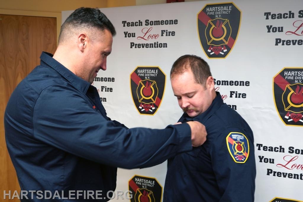 Two firefighters are performing a ceremonial act, possibly pinning a badge, against a backdrop promoting fire prevention.