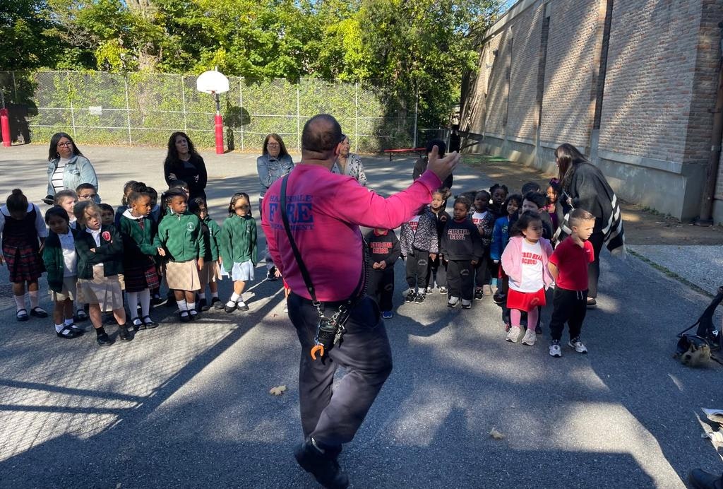 A group of young children in school outfits listens to an adult in pink, possibly during an outdoor educational event.