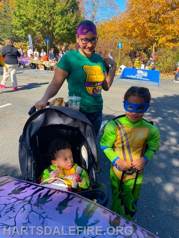 A woman and two children, one in a stroller and one in a superhero costume, enjoy a festive outdoor event on a sunny day.