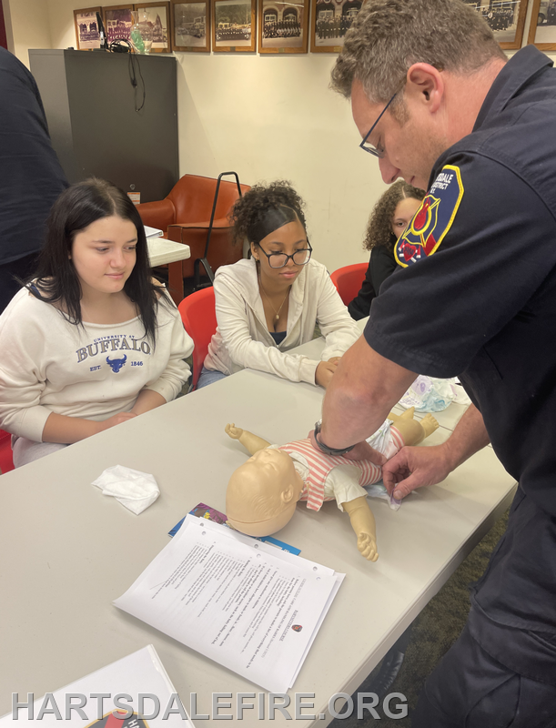A firefighter teaches CPR using a doll while a few students observe and learn at a classroom setting.