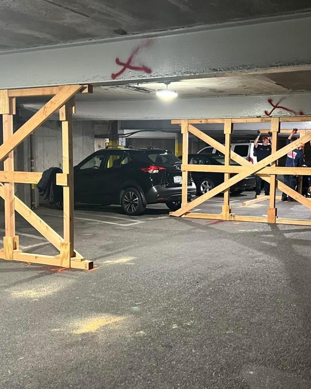 The image shows a parking garage with wooden barriers blocking off a section, and people nearby, possibly inspecting or managing the area.