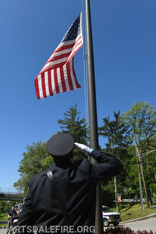 A person in uniform saluting the U.S. flag raised on a pole under a clear blue sky.