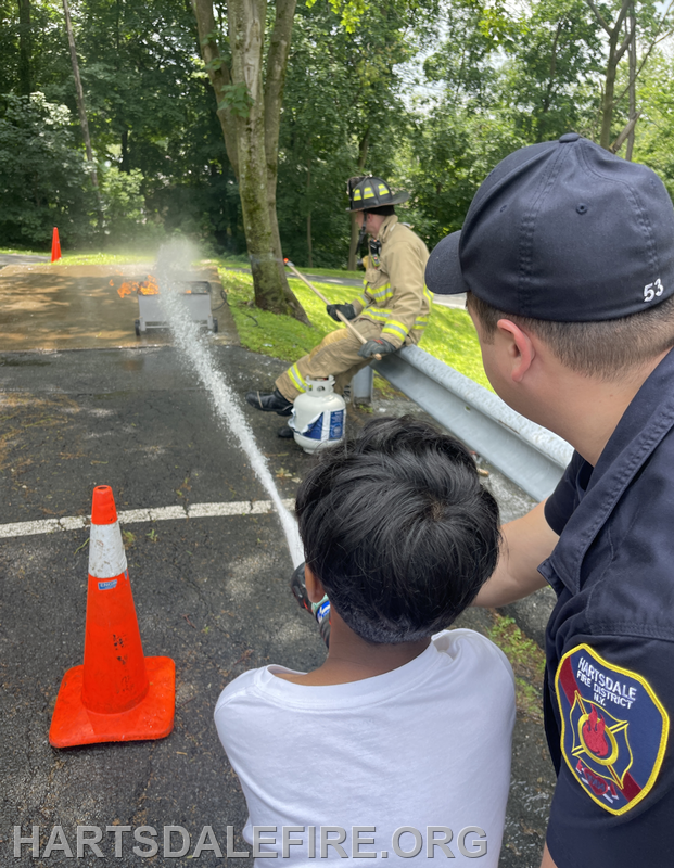 A child practices using a fire extinguisher under a firefighter’s guidance, while a small controlled fire is present.