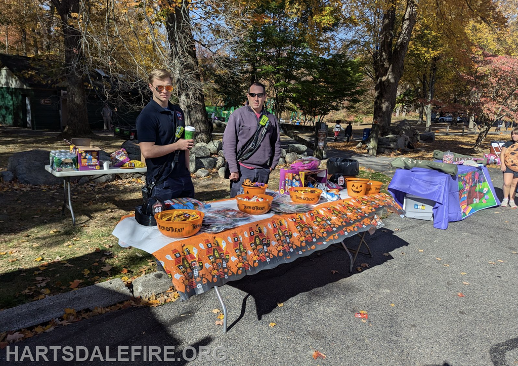 Two people at a fall event stand behind a table with Halloween-themed treats and decorations. There are snacks and colorful displays.