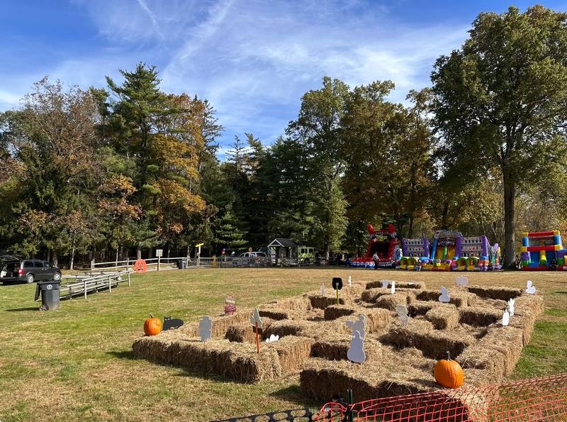An autumn scene with hay bales arranged in a maze, pumpkins, and bounce houses in the background, surrounded by trees.