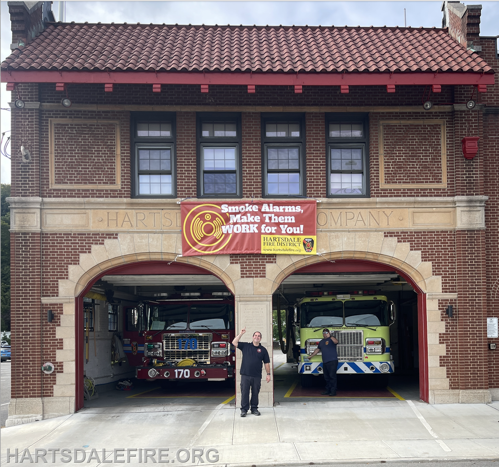 A historic fire station with two fire trucks and a banner promoting smoke alarm safety, plus two firefighters waving.