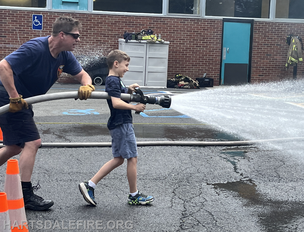 A firefighter helps a boy operate a fire hose, joyfully spraying water during a fire safety event.