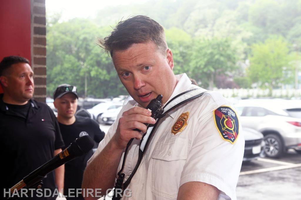 A fire department official speaks into a microphone, while others listen nearby in an outdoor setting.