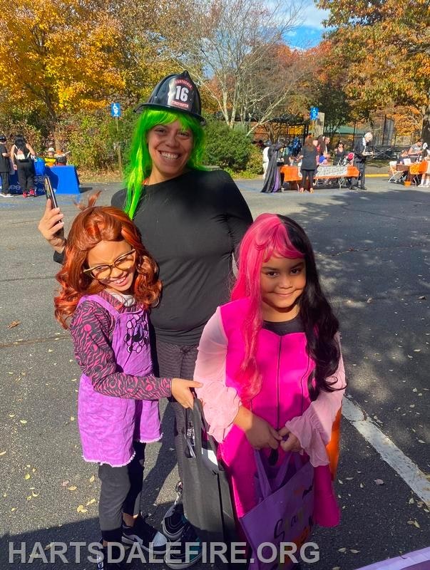 A woman in a firefighter hat with green hair poses with two children in fun costumes and colorful wigs at an outdoor event.
