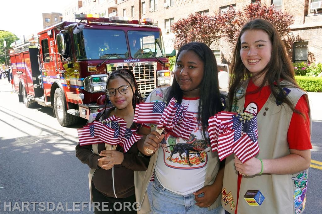Three girls holding small American flags pose in front of a fire truck during a community event.
