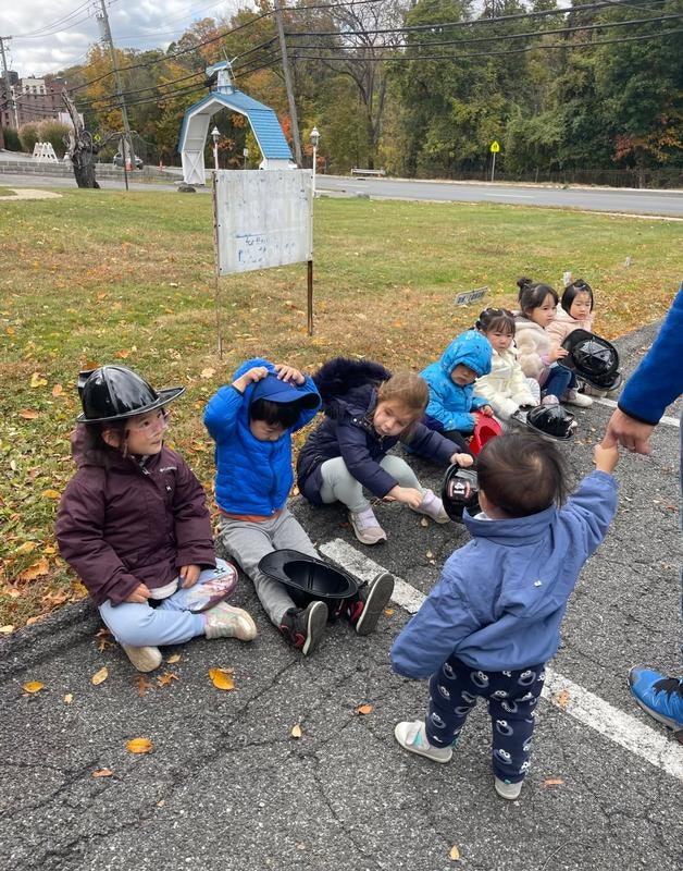 A group of children sits on the ground, wearing jackets and helmets, in a chilly outdoor setting, interacting with each other.