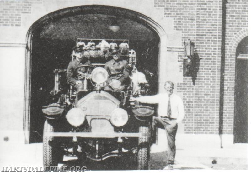 Vintage fire truck with firefighters and a man standing beside it, in front of a brick building.