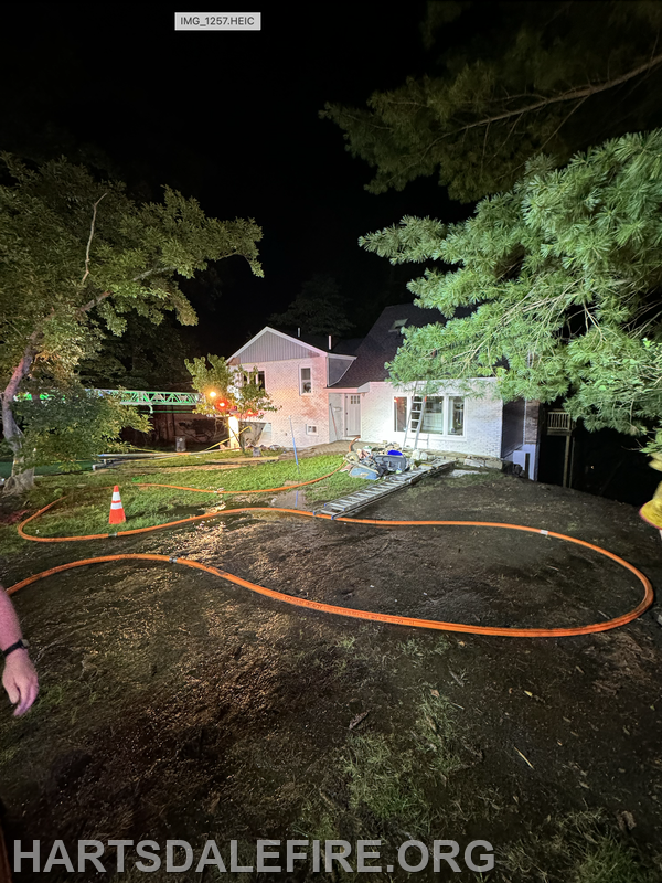 A nighttime scene showing a house, fire equipment, hoses, and emergency lights, indicating a fire response situation.