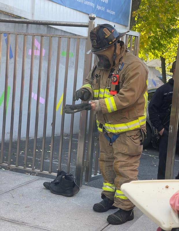 A firefighter in gear is using a tool near a gate, with a pair of boots on the ground beside him.