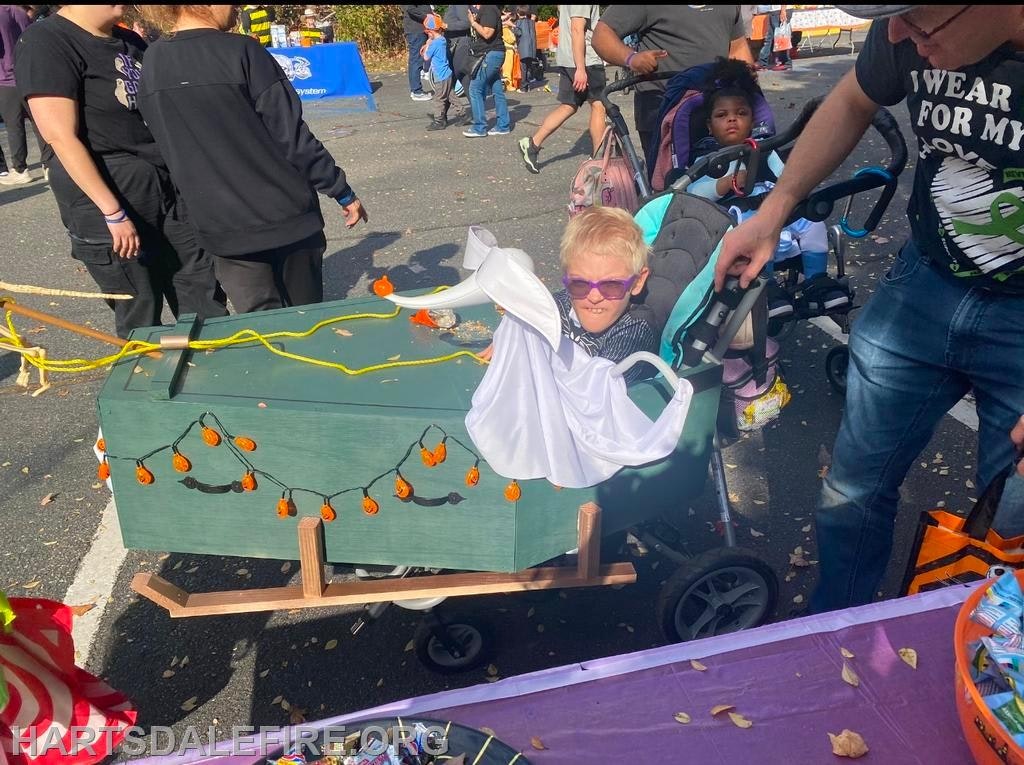 A child in sunglasses sits in a decorated cart resembling a coffin, at a festive outdoor event with people around.
