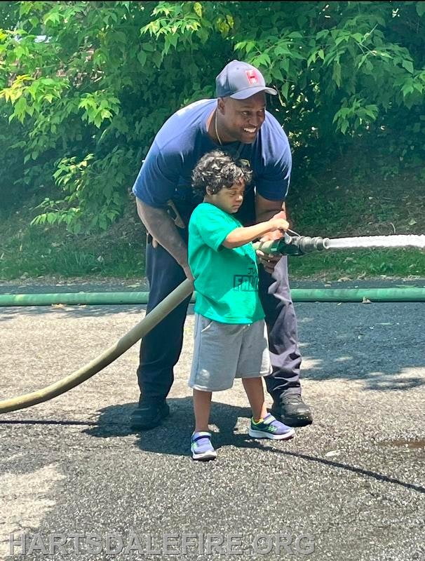 A firefighter helps a child hold a fire hose, spraying water on a sunny day.