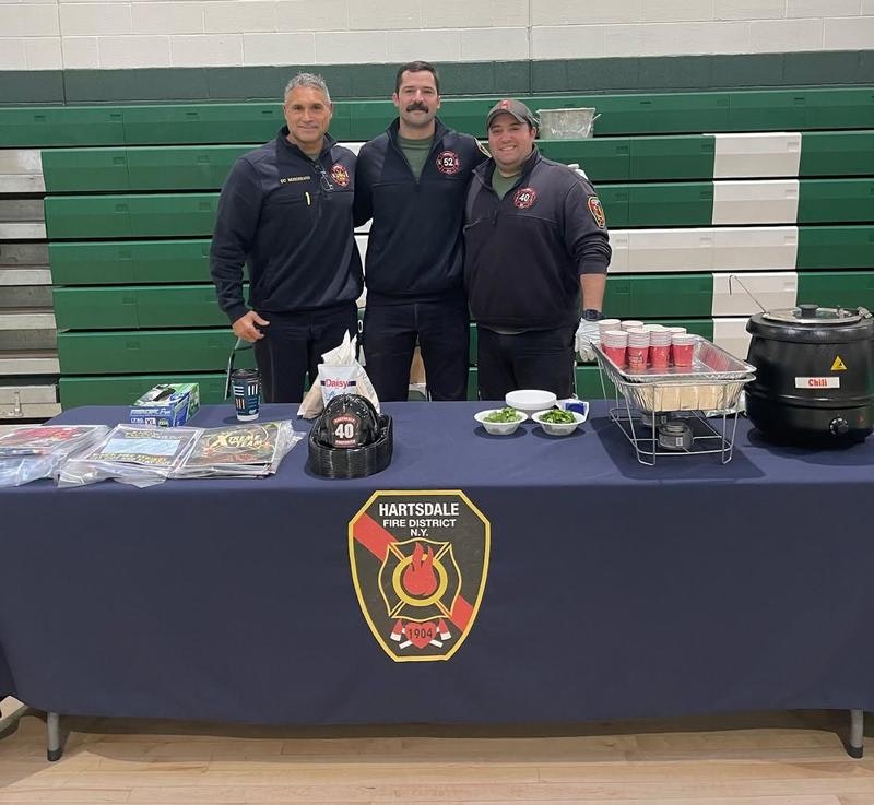Three firefighters from Hartsdale Fire District stand behind a table with food and promotional materials in a gymnasium.