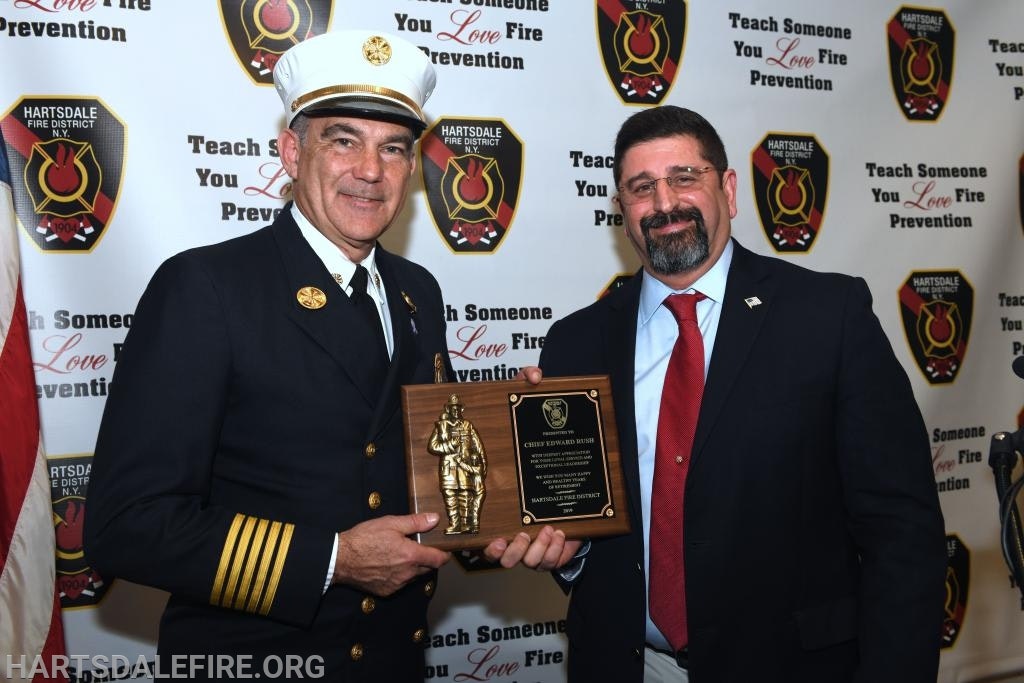 Two men stand holding an award plaque against a backdrop promoting fire prevention.