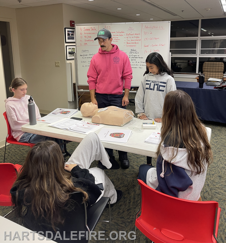 A group of young people learning CPR techniques with a trainer and a practice dummy in a classroom setting.