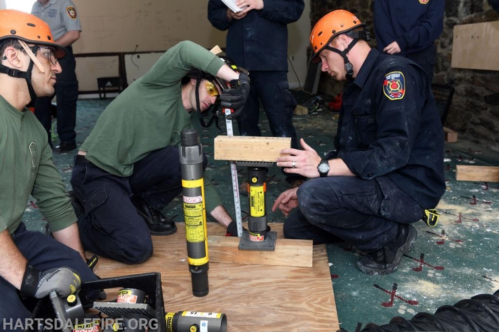 People are wearing helmets, measuring, and working with tools indoors, possibly for a construction or safety training.