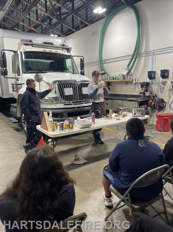 Two presenters are demonstrating safety or cooking tips at a table in a fire station, with an audience and a fire truck in the background.