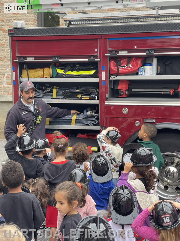 A firefighter shows equipment to a group of children wearing helmets near a fire truck.