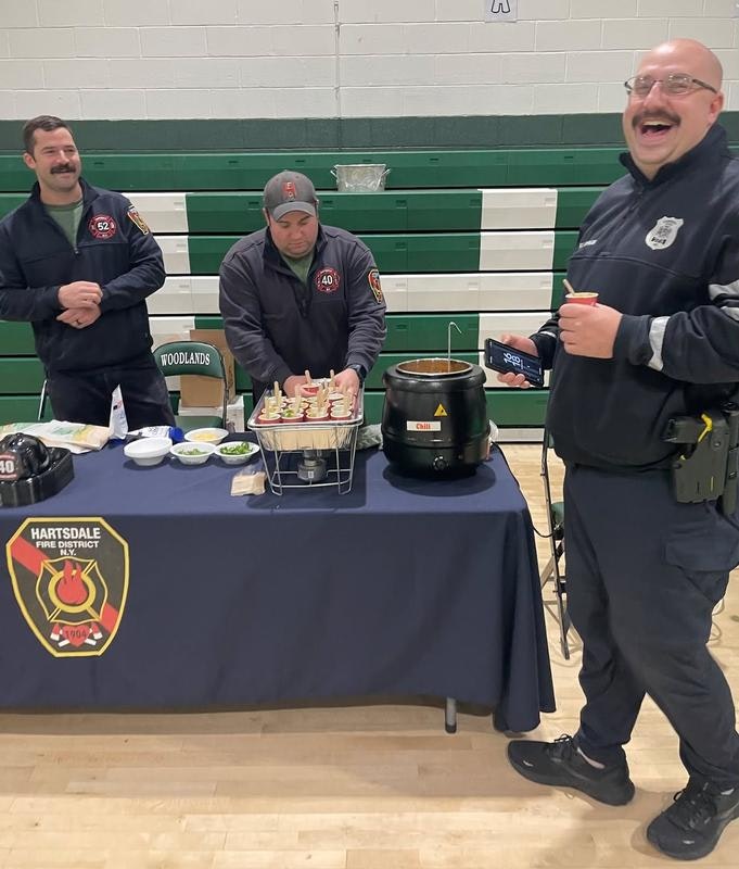Firefighters and a police officer are enjoying a cheerful moment at a table with food, including chili, in a gymnasium.