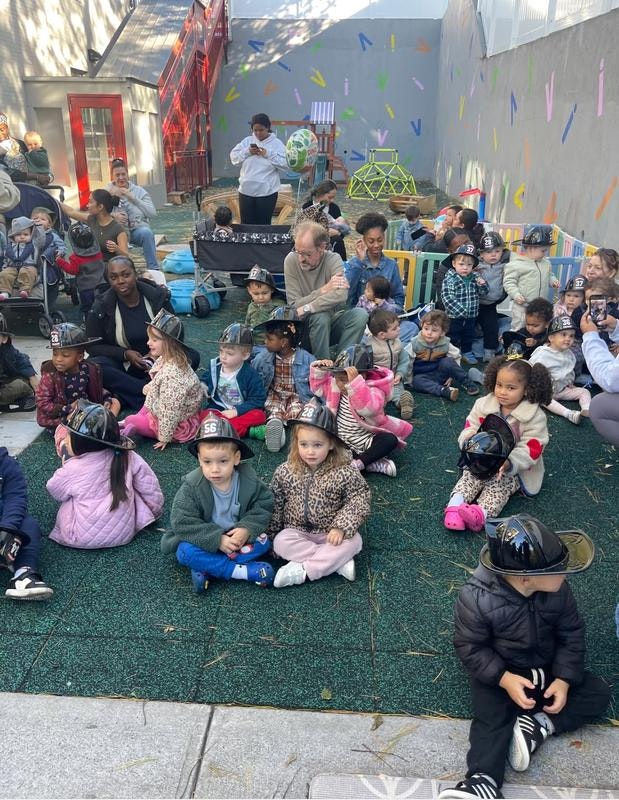 A group of children wearing hats sits on a grassy area, with adults nearby in a playful, colorful outdoor setting.